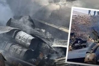Wreckage from Iranian Army helicopter crash in Isfahan showing twisted metal debris, oil drums, and damaged fuselage amid smoke.