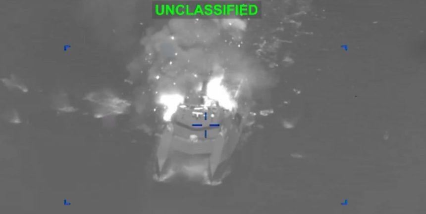 A U.S. Navy warship on open water engaging a small hostile vessel near the Strait of Hormuz, with smoke rising from the target area under a clear sky.