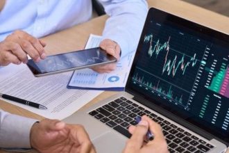 Two people sit at a desk with a laptop showing stock market charts and a tablet displaying financial data, alongside printed documents and a pen.