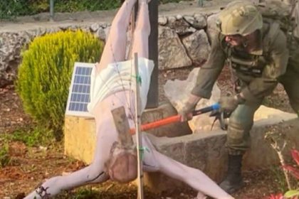 A uniformed IDF soldier uses a sledgehammer to smash Jesus Statue in a garden-like in southern Lebanon setting bordered by stone walls and shrubs.
