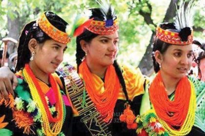 Three Kalash women in colourful traditional clothing, beaded necklaces, and feathered headpieces pose together outdoors.