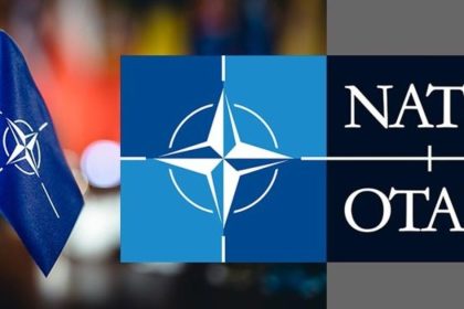 Blue NATO flag beside the NATO/OTAN emblem on a blurred multicolored background.