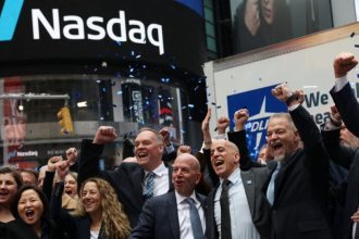 Group of people cheering with raised fists and confetti beneath a large Nasdaq sign.