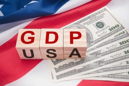 Wooden letter blocks spelling GDP and USA on top of US hundred-dollar bills and an American flag.