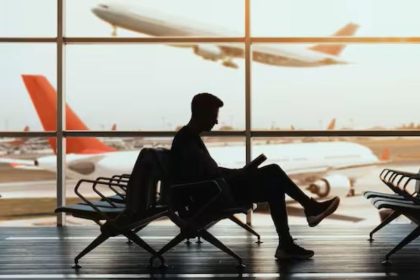 A silhouetted traveler sits in an airport terminal, reading or using a device near large windows overlooking planes on the runway.