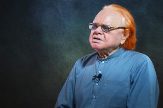 Veteran journalist Altaf Hassan Qureshi wears glasses and a blue kurta while seated against a dark studio background.