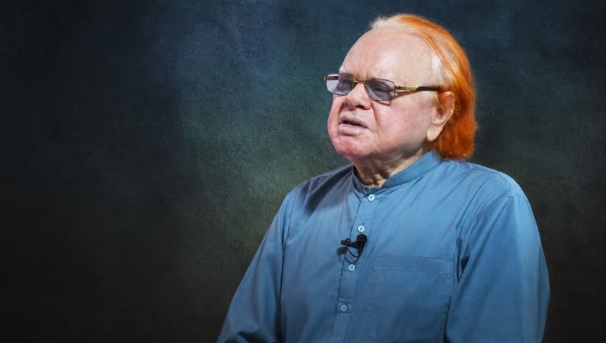 Veteran journalist Altaf Hassan Qureshi wears glasses and a blue kurta while seated against a dark studio background.