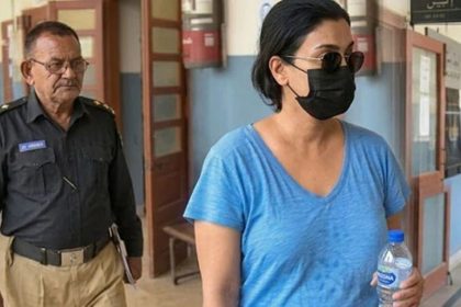 Anmol Pinky walks through a courthouse hallway wearing sunglasses and a black face mask, while a police officer follows behind her.