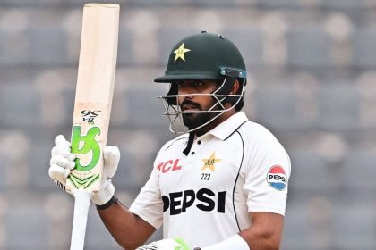 Pakistan cricketer Babar Azam raises his bat while wearing a white Test uniform and green helmet.
