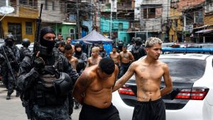 A Brazilian law enforcement officer in tactical gear escorts two shirtless suspected gang members near a police vehicle during an urban operation.