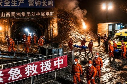 Emergency workers in orange uniforms carry stretchers outside a coal mine entrance at night in Shanxi, China.