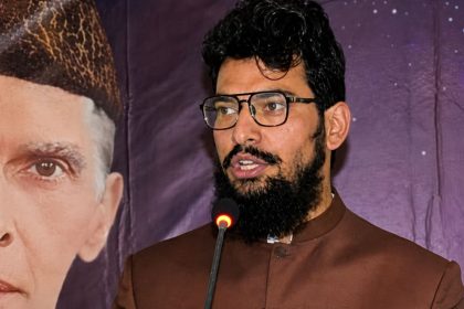 Hamza Ahmed Dar ins glasses and a brown traditional outfit speaks at a microphone beside a large portrait of Muhammad Ali Jinnah.