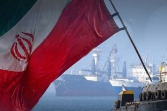 Iranian flag waving in front of ships and port cranes near a waterfront.