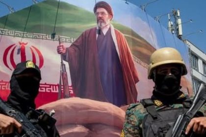 Two armed Iranian security personnel stand before a large banner showing Iran’s Supreme Leader Mojtaba Khamenei and the Iranian flag.