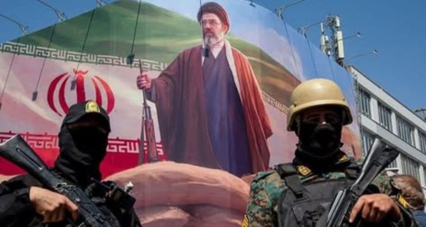 Two armed Iranian security personnel stand before a large banner showing Iran’s Supreme Leader Mojtaba Khamenei and the Iranian flag.