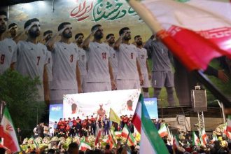 Crowds wave Iranian flags at night in front of a large banner showing Iran’s national football team players standing in uniform.