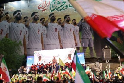 Crowds wave Iranian flags at night in front of a large banner showing Iran’s national football team players standing in uniform.