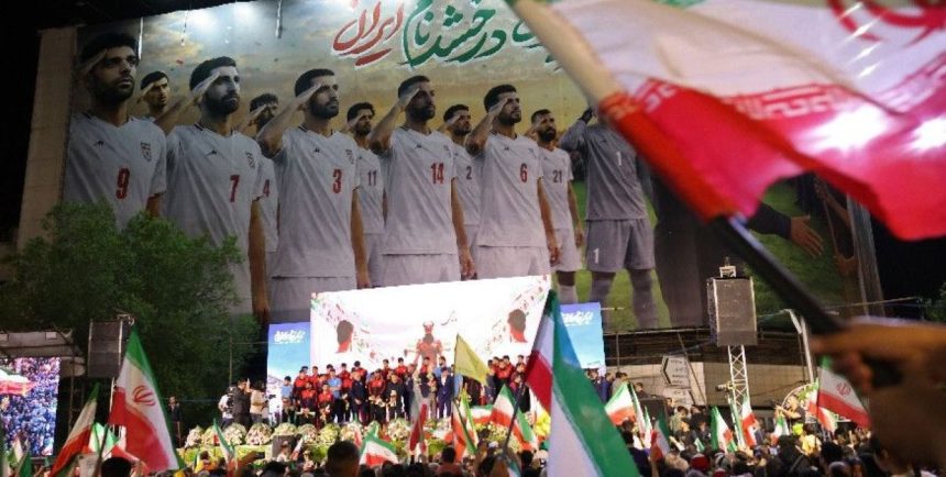 Crowds wave Iranian flags at night in front of a large banner showing Iran’s national football team players standing in uniform.