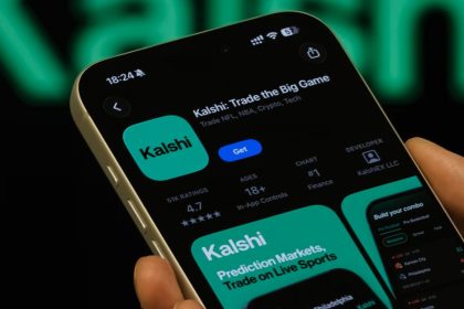 A hand holds a smartphone showing the Kalshi app page, with prediction market and live sports trading graphics on the screen.
