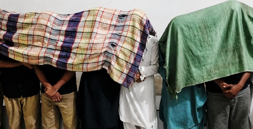 Suspects arrested by Karachi Police in a kidnapping case stand against a wall with cloths covering their faces.