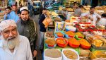 A busy Karachi market shows vendors and shoppers standing beside colorful bowls of spices, sacks of grains, dried fruits and packaged goods.