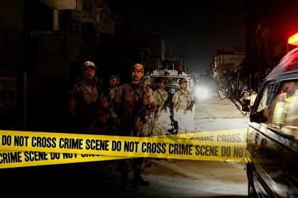 Security personnel guard a nighttime street behind yellow crime scene tape, with an emergency vehicle on the side