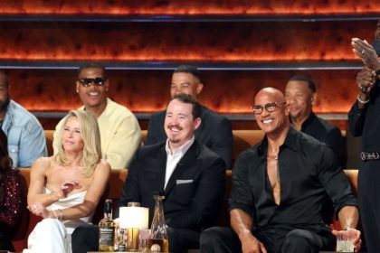 Kevin Hart stands with a microphone during a Netflix roast event, with seated celebrity guests and performers smiling nearby.