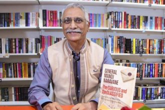 Former Indian Army Chief General (retd.) Manoj Naravane sits in front of bookshelves while holding his book, The Curious and the Classified.