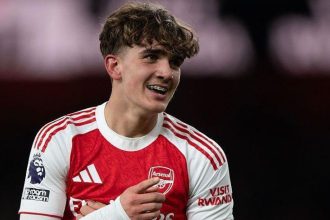 Max Dowman smiles while wearing an Arsenal home shirt on the pitch.