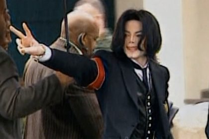 Michael Jackson wears a dark suit and gestures with one hand while standing among security and other people.
