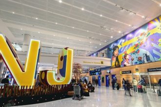 ALT Text: Newark Liberty International Airport’s Terminal A interior shows a large NJ display, travelers and colorful wall artwork.