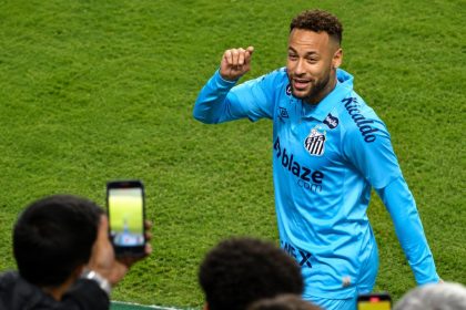 Neymar stands on a football pitch in a light blue Santos kit as fans hold up phones near the sideline.