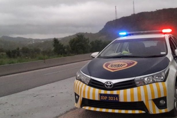 A Pakistan Motorway Police car with flashing red and blue lights stands on the side of a highway near green hills under cloudy skies.