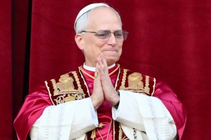 Pope Leo XIV wears red and white papal vestments while standing before a red curtain at St. Peter’s Basilica.