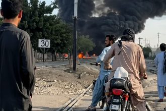 People on motorcycles watch thick black smoke rising near a railway track after a reported train blast near Chaman Phatak in Quetta.