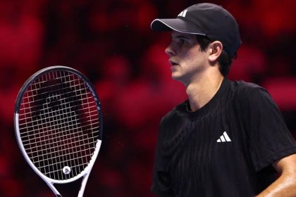 Tennis player Rafael Jodar, wearing a black Adidas shirt and cap, holds a racket during a match against a blurred red background.