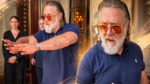 Russell Crowe gestures outside a hotel while holding a pen, with staff visible nearby. Image Credit: Screengrab