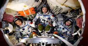 Three Shenzhou-23 astronauts sit inside a compact spacecraft module while wearing white spacesuits with blue trim.