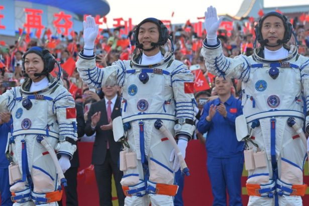 Three Chinese astronauts in white spacesuits wave to supporters holding flags before the Shenzhou-23 mission.