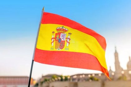 The flag of Spain waves in the foreground with a sunlit, blurred city skyline in the background.