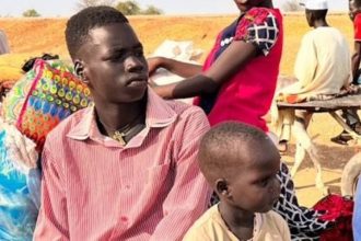 A young man sits with children and other people in a dry outdoor area, with bags and belongings nearby during Sudan’s hunger crisis.