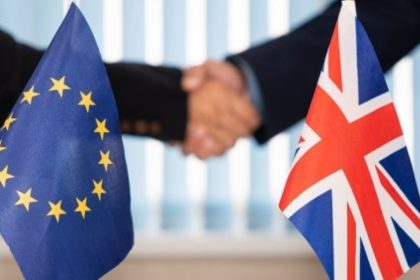 Small EU and UK flags stand in front of two people shaking hands during a diplomatic meeting.