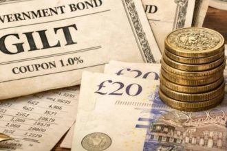 A UK government bond certificate labeled “Gilt” sits beside £20 notes, stacked coins, a calculator and financial papers.
