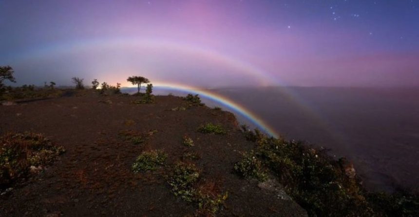 A moonbow glows over Victoria Falls under a purple night sky, with mist, stars and rugged ground in the foreground.