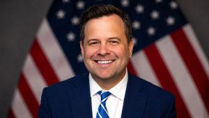 Bill Pulte smiling in a suit with an American flag in the background.