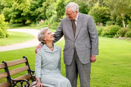 The Duchess of Gloucester sits on a garden bench while The Duke of Gloucester stands beside her in the gardens of Buckingham Palace.