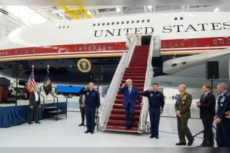 Donald Trump walks down red-carpeted aircraft stairs beside a large United States government plane inside a hangar.