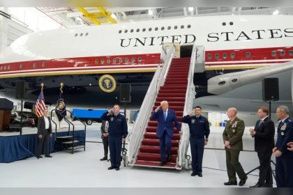 Donald Trump walks down red-carpeted aircraft stairs beside a large United States government plane inside a hangar.