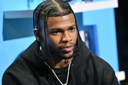 Shilo Sanders sits in a black hoodie during an interview-style appearance against a blue backdrop.