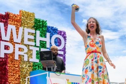 A person in a colorful dress raises one arm beside a rainbow “WeHo Pride” sign at an outdoor Pride event.